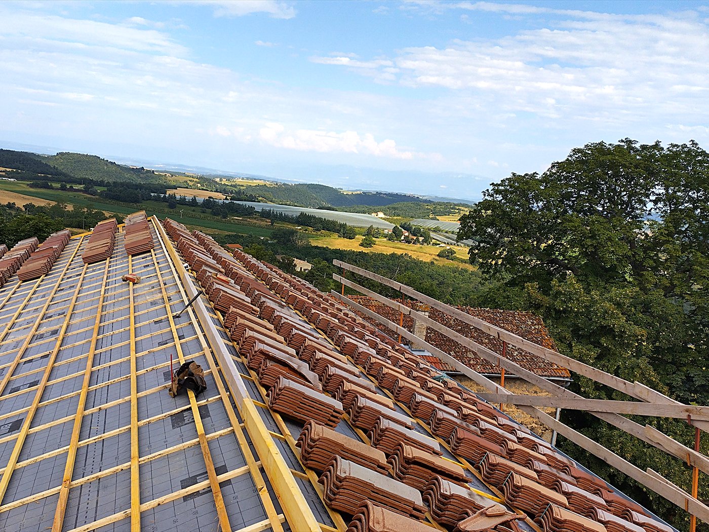 Couvreur posant les tuiles sur les liteaux d&rsquo;un grand pan de toiture, avec vue sur la campagne ardéchoise